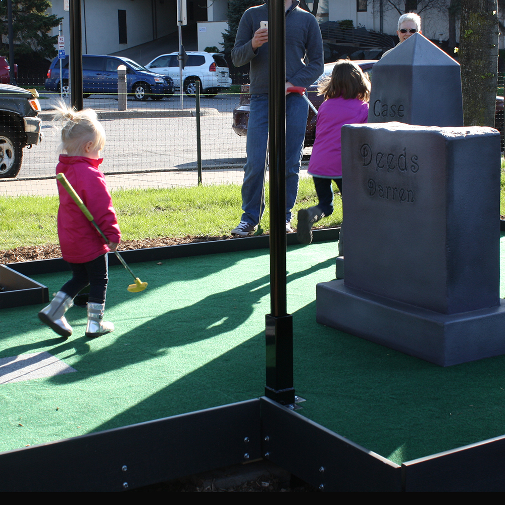 Mini-golf hole design to look like a cemetery, with steel gate, fiberglass tombstones, and artificial turf.